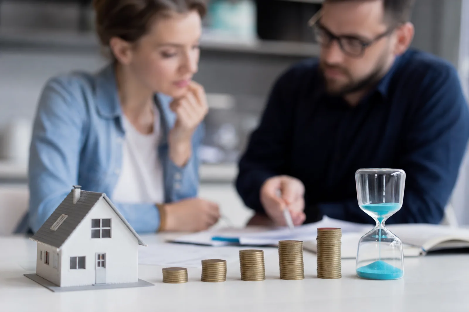 Couple examinant des plans de financement immobilier à table, avec une maison miniature et des documents bancaires.
