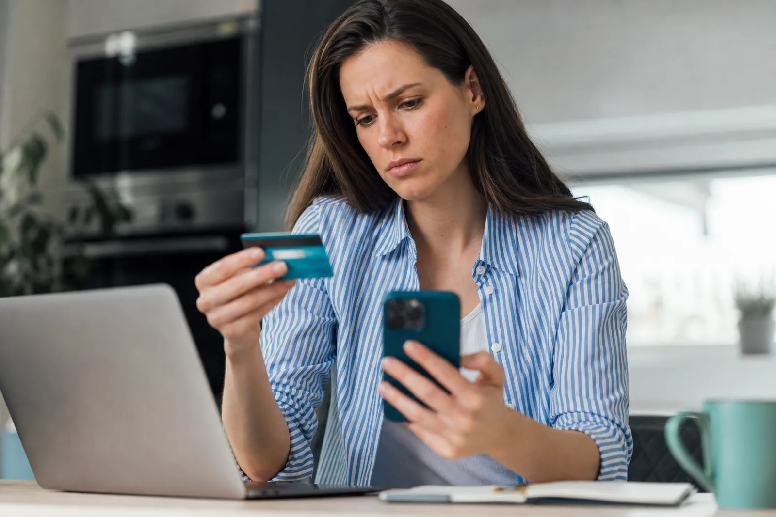 Une femme inquiète tient sa carte bancaire et son téléphone devant un ordinateur portable, en train de vérifier une opération suspecte.