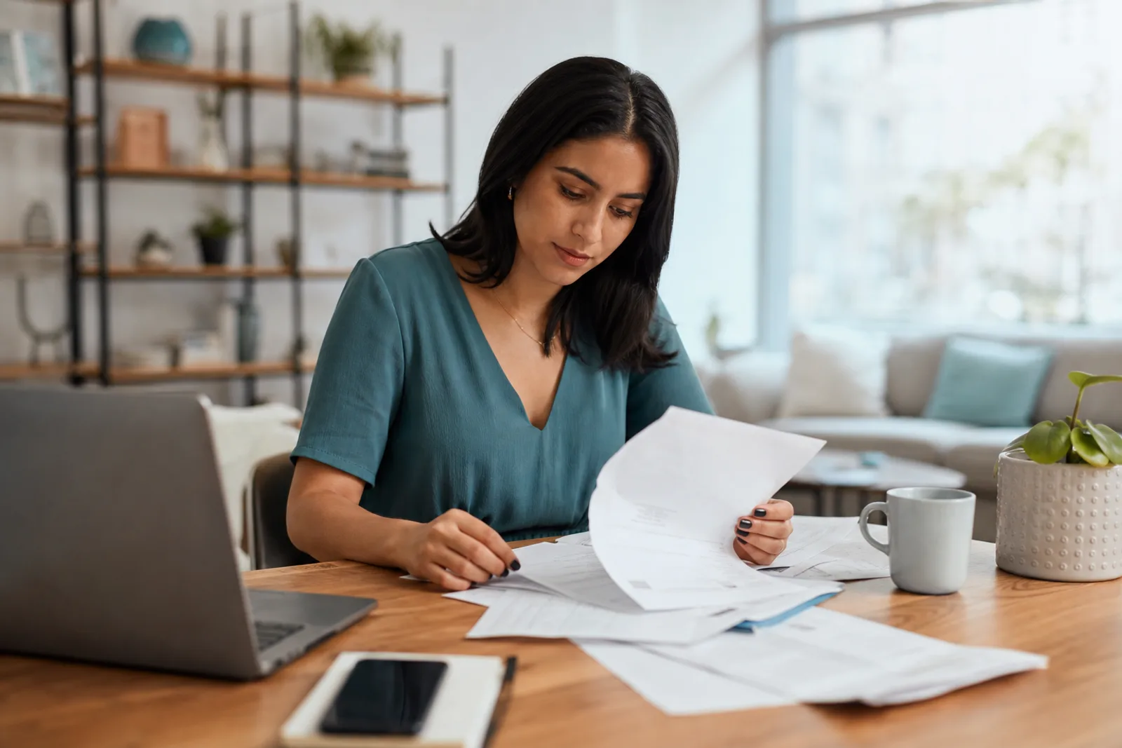 Une femme trie des relevés bancaires étalés sur une table, devant un ordinateur portable, dans un salon lumineux.